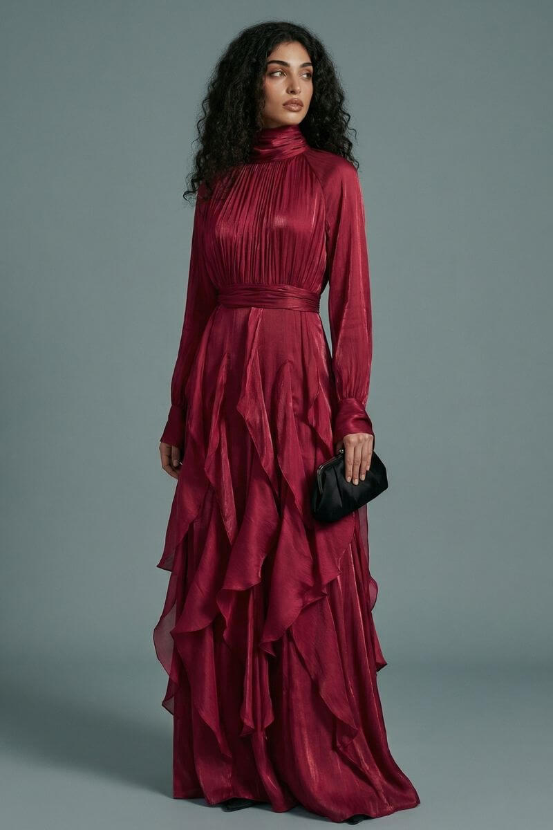 A model in a burgundy high-neck maxi dress is shown in a three-quarter pose, highlighting the fluid drape, gathered waist, and dimensional ruffle detailing.