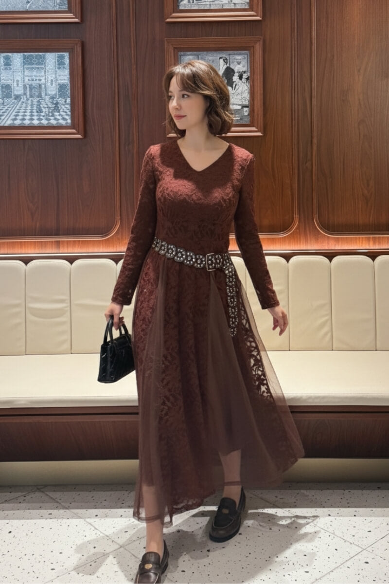 Woman wearing a burgundy lace and mesh midi dress styled with a jeweled belt, holding a black handbag and loafers, posing in an elegant wood-paneled lounge.
