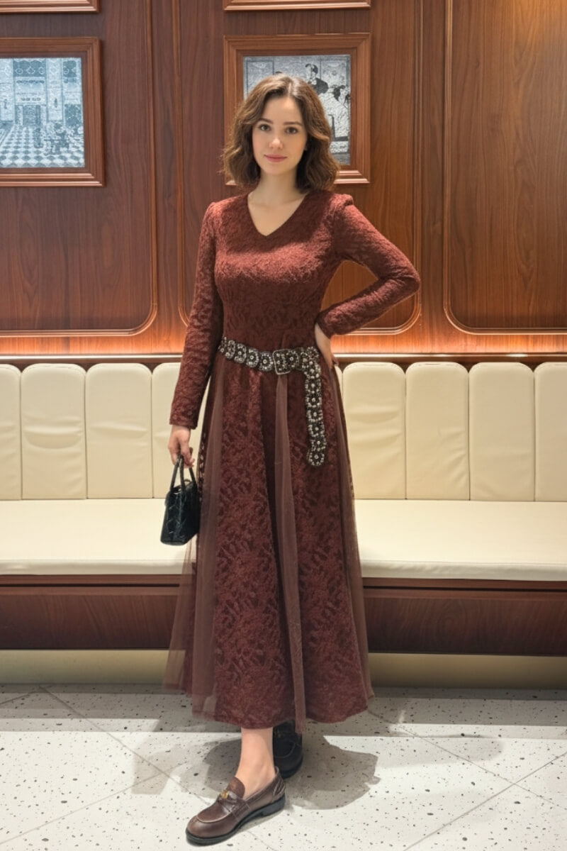 Woman standing in a sophisticated interior wearing a burgundy lace mesh midi dress with a V-neckline and beaded belt, paired with brown loafers and a black handbag.