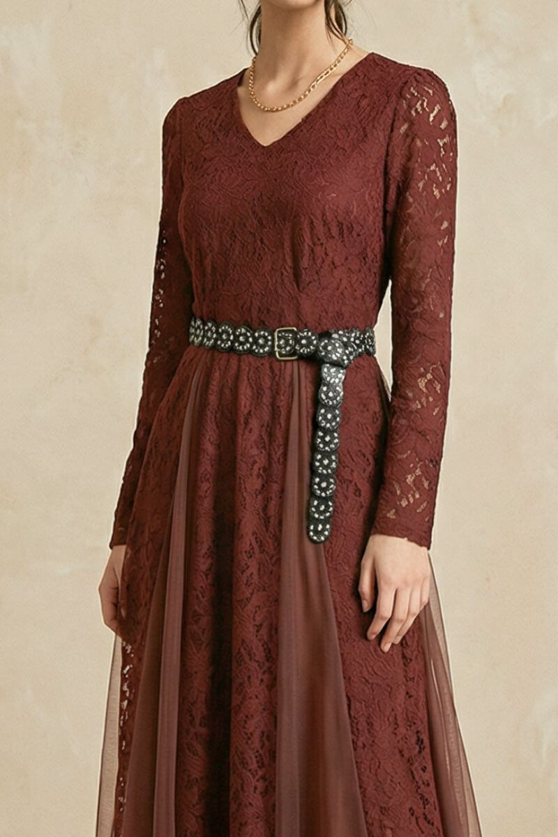 A model wears a burgundy lace V-neck midi dress with sheer long sleeves, a decorative studded belt, and a softly layered skirt against a warm neutral studio backdrop.