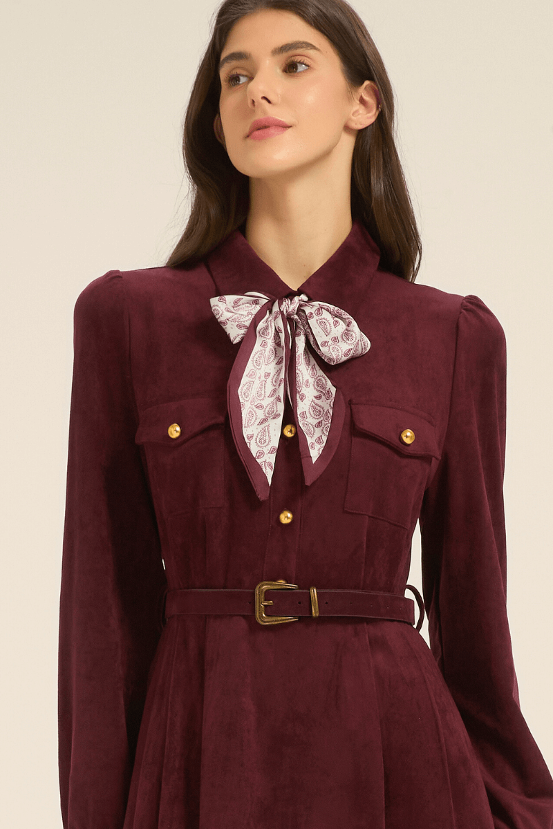 Close-up of a burgundy midi dress showing the collared neckline, gold buttons, chest pockets, belted waist, and a patterned bow scarf tied at the neck.