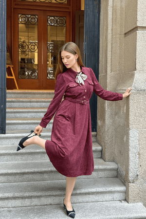 Model in a burgundy belted midi dress with chest pockets and a bow tie scarf, striking a playful pose by lifting one heel.