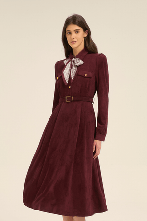 Model in a burgundy midi dress styled with a slim belt and paisley bow scarf at the collar, standing in a relaxed angled pose.