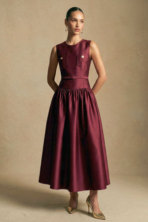 Model poses in a deep burgundy tailored midi dress featuring a sleeveless cut, defined waist belt, and fluid A-line skirt, photographed in a neutral studio setting.