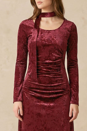 A close-up of a model wearing a burgundy velvet midi dress with a soft scoop neckline, long fitted sleeves, subtle ruching at the waist, and an integrated velvet choker scarf detail against a warm neutral studio backdrop.