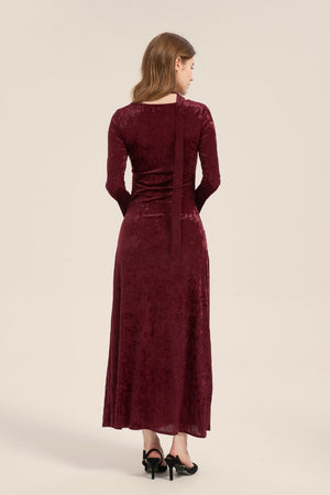 Model wearing a burgundy velvet maxi dress with long sleeves and scarf tie neckline, fitted silhouette and soft sheen, styled with black heels.