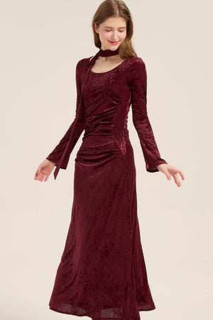 Model turning slightly in a burgundy velvet long dress, showing soft draping and slim silhouette with tie scarf detail at neck.