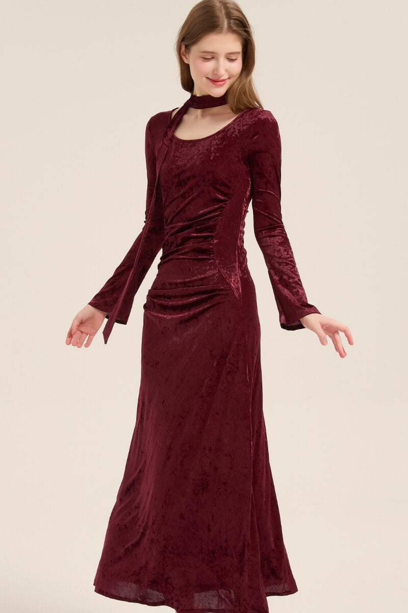 Model turning slightly in a burgundy velvet long dress, showing soft draping and slim silhouette with tie scarf detail at neck.