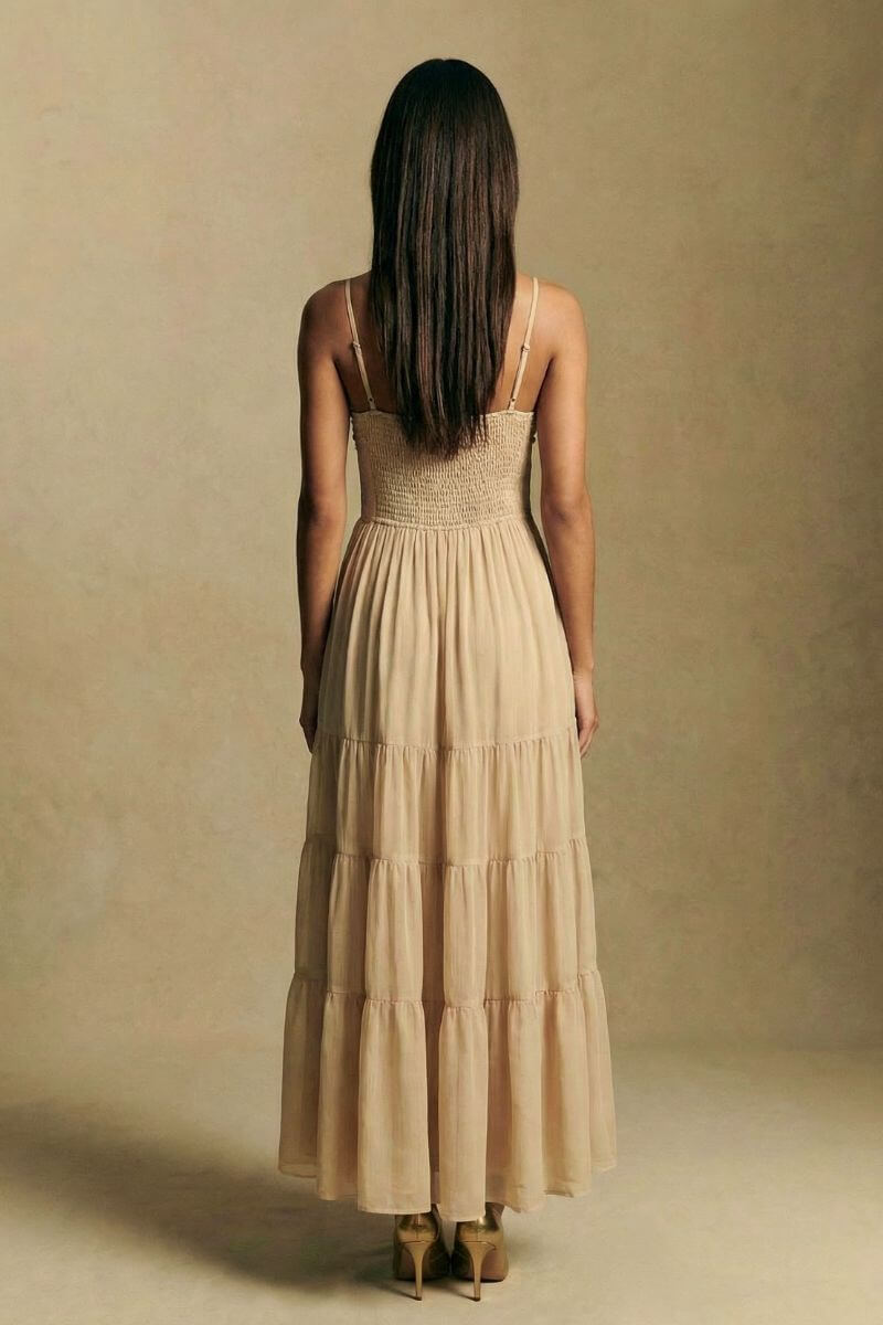 Back view of a champagne embroidered maxi dress featuring a smocked bodice, adjustable spaghetti straps, and a softly tiered skirt, photographed against a minimal beige backdrop.