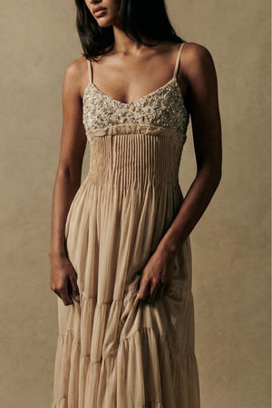 A close-up front view of a model wearing a champagne-toned maxi dress with delicate embroidered bodice detailing, soft pleated waist, and flowing tiered chiffon skirt, styled in a warm neutral studio setting.