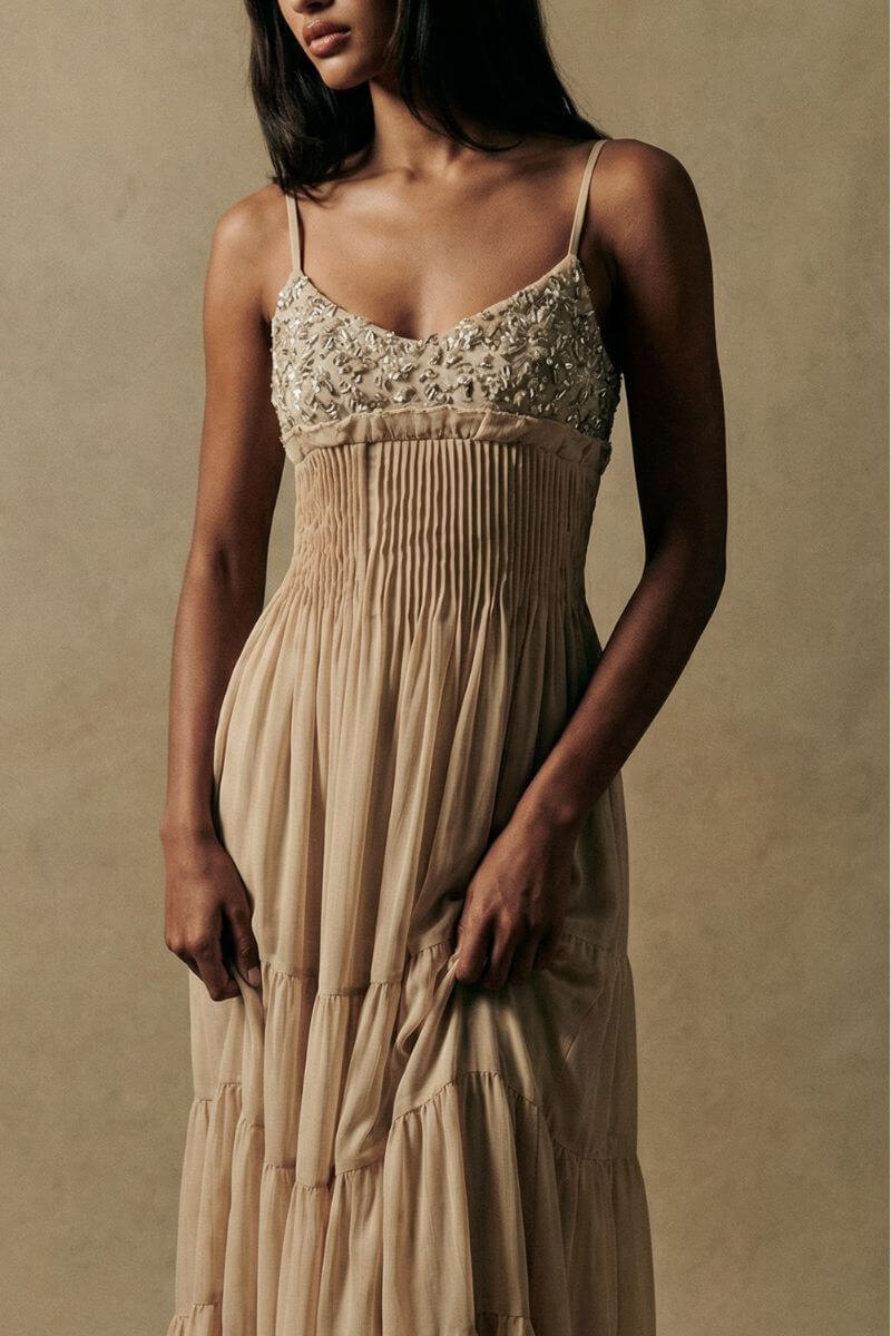A close-up front view of a model wearing a champagne-toned maxi dress with delicate embroidered bodice detailing, soft pleated waist, and flowing tiered chiffon skirt, styled in a warm neutral studio setting.