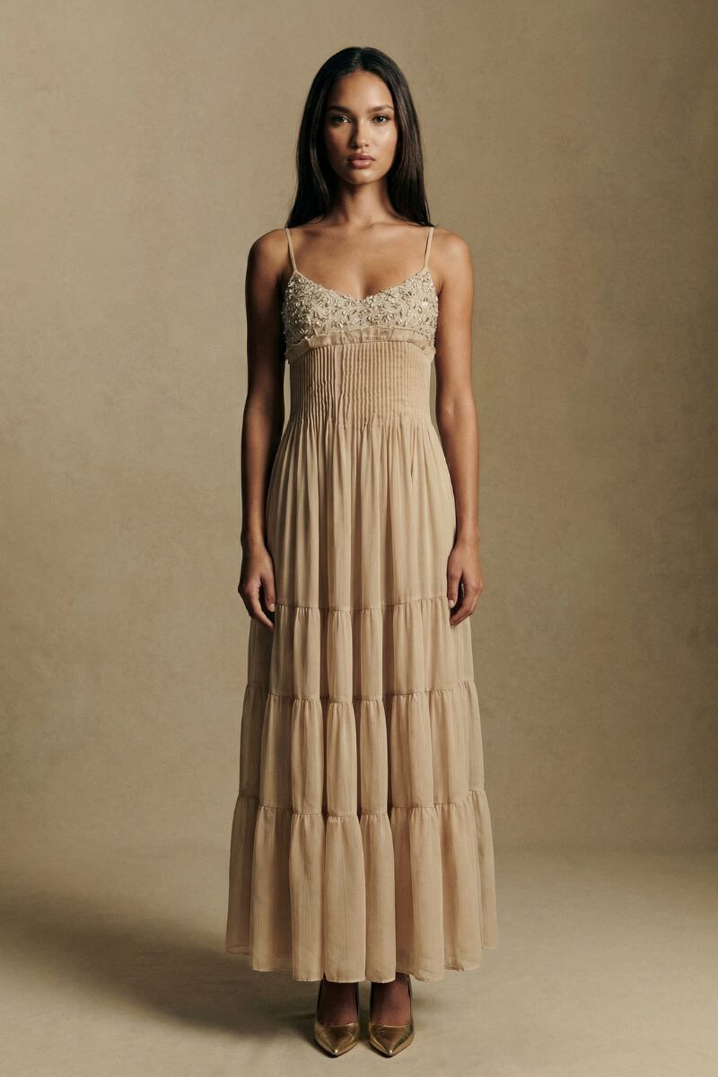 A full-length front view of a model wearing a champagne tiered maxi dress with an embroidered bust, pleated waistline, and airy chiffon layers, paired with pointed heels in a refined studio setting.