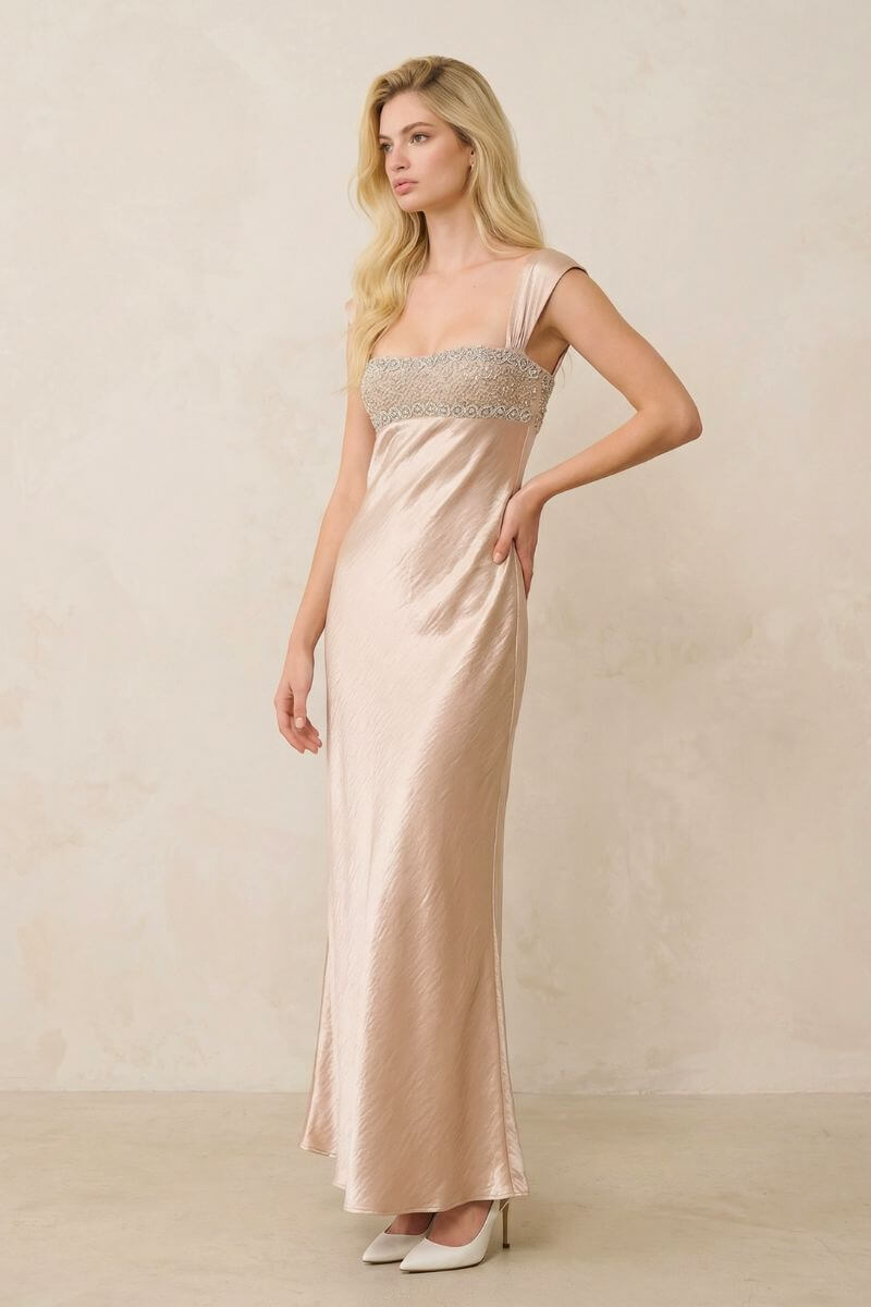 A blonde model poses in a champagne satin maxi dress featuring a beaded bust panel and wide straps, styled with ivory heels in a minimalist studio setting.