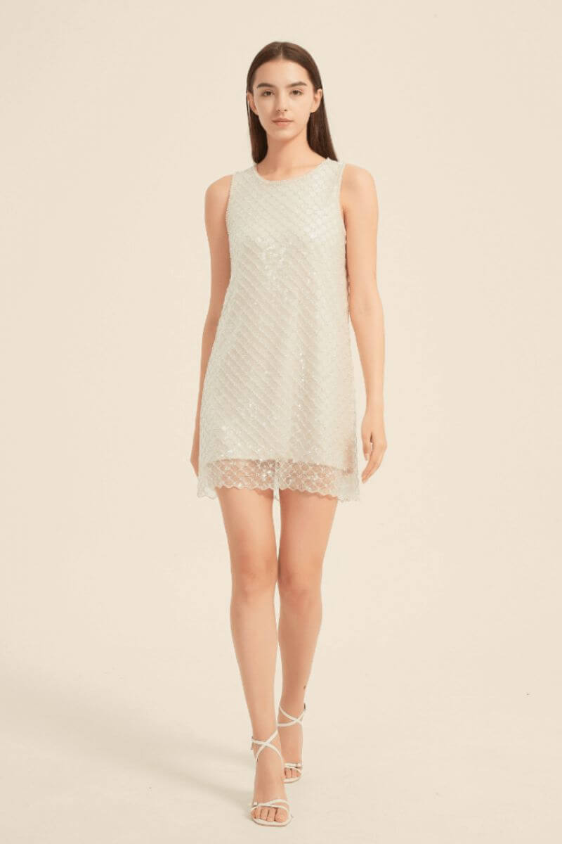 Woman wearing a sleeveless champagne sequin mini dress with scalloped hem and diamond-pattern shimmer, styled with white heels.