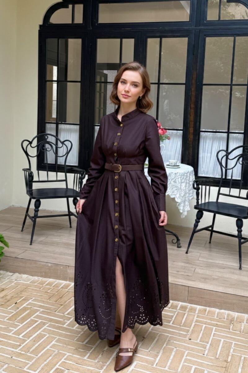 Model standing on a tiled terrace wearing a chocolate brown button-down maxi shirt dress with a belted waist, mandarin collar, and scalloped eyelet hem, paired with brown pointed-toe flats near black wrought-iron chairs and a lace-covered café table.