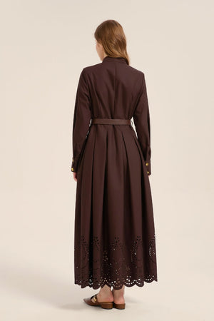 Model turned to show the back of a chocolate brown cotton maxi dress featuring a belted waist, long sleeves, and laser-cut eyelet hem detail.