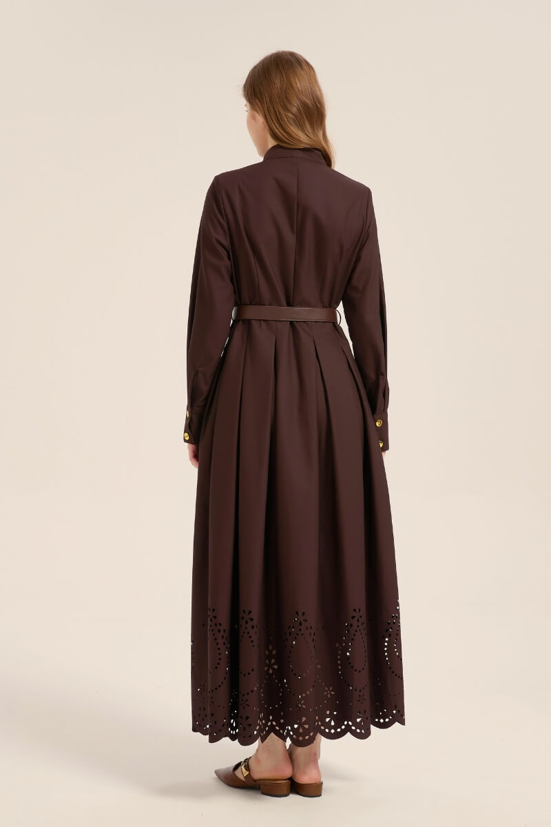 Model turned to show the back of a chocolate brown cotton maxi dress featuring a belted waist, long sleeves, and laser-cut eyelet hem detail.