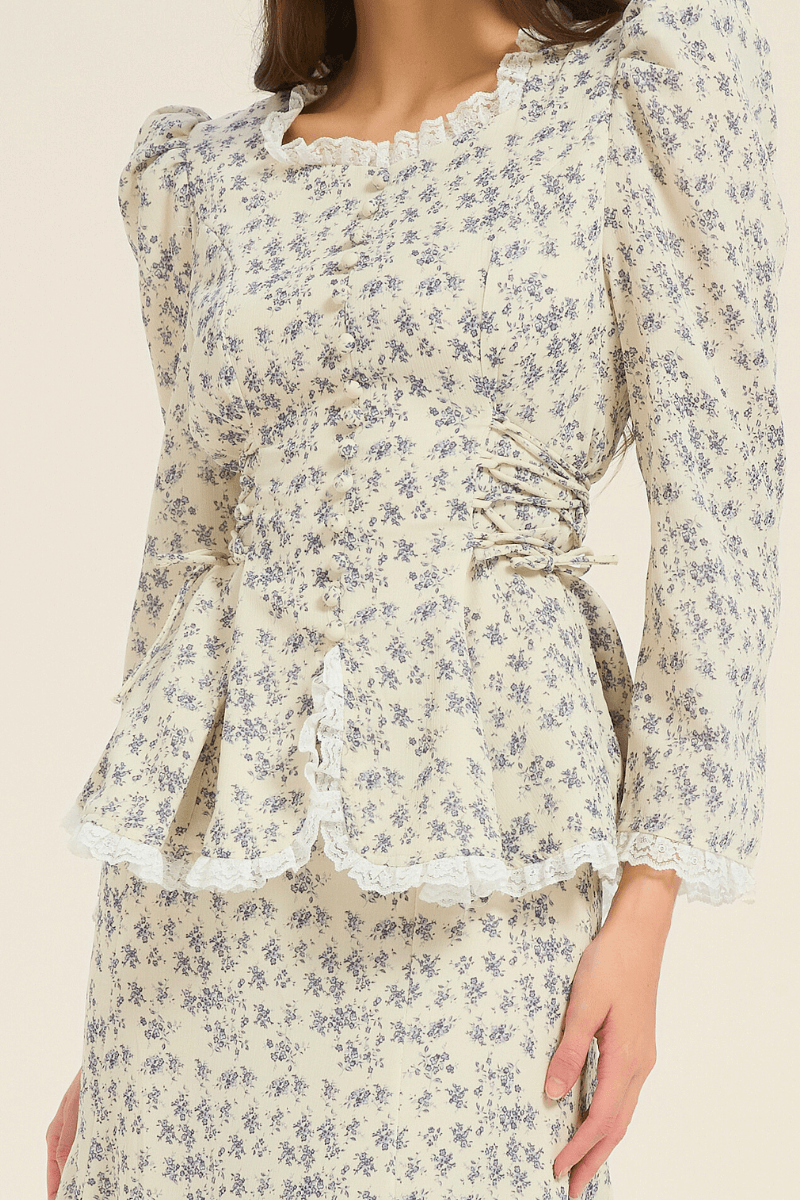 Close-up of the cream and blue floral peplum blouse showing fabric-covered buttons, lace trim, and side-tie detail.