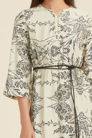 Detailed close-up showing the notch neckline, covered buttons, and black waist tie on a cream midi dress with intricate botanical print.