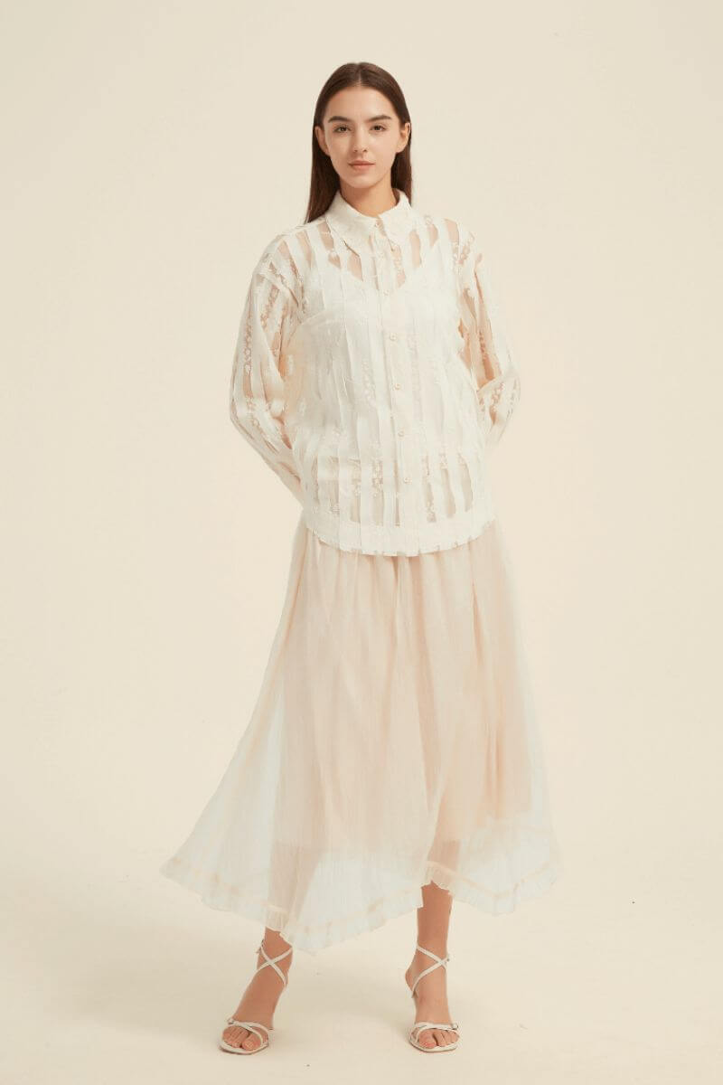 Close-up shot of a blush ivory midi skirt with sheer layered fabric and delicate ruffled hem, styled with white heeled sandals.