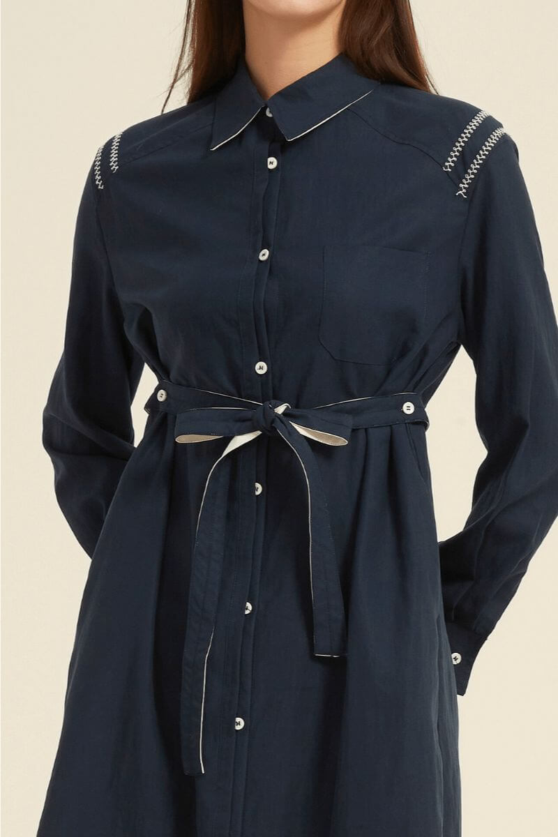 Detailed close-up of a navy button-down midi shirt dress featuring white contrast stitching on the shoulders and a belted waist tie — minimalist tailoring by Sioéne.