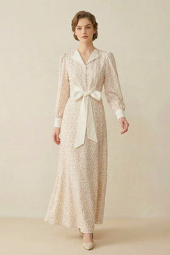 A woman in a cream floral maxi dress with a satin collar, long sleeves, and a large front bow detail, standing in a minimal studio setting.