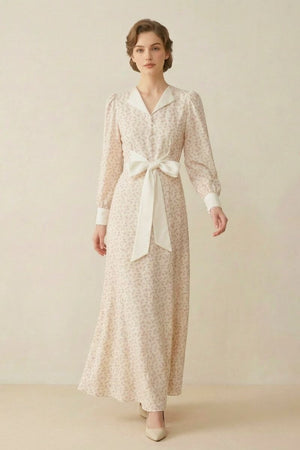 A woman in a cream floral maxi dress with a satin collar, long sleeves, and a large front bow detail, standing in a minimal studio setting.