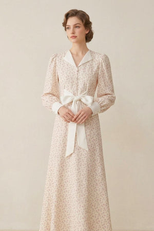 A model wearing a cream floral print maxi dress featuring a satin notched collar, puff sleeves, and a long front bow, posed in a soft neutral studio.