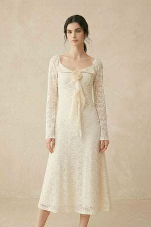 A woman standing in a cream lace long-sleeve midi dress featuring a sweetheart neckline and a fabric flower detail with a trailing ribbon.