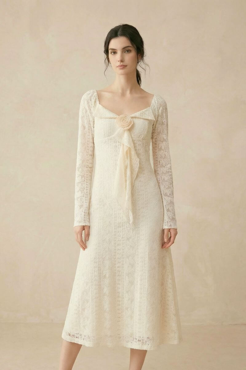 A woman standing in a cream lace long-sleeve midi dress featuring a sweetheart neckline and a fabric flower detail with a trailing ribbon.