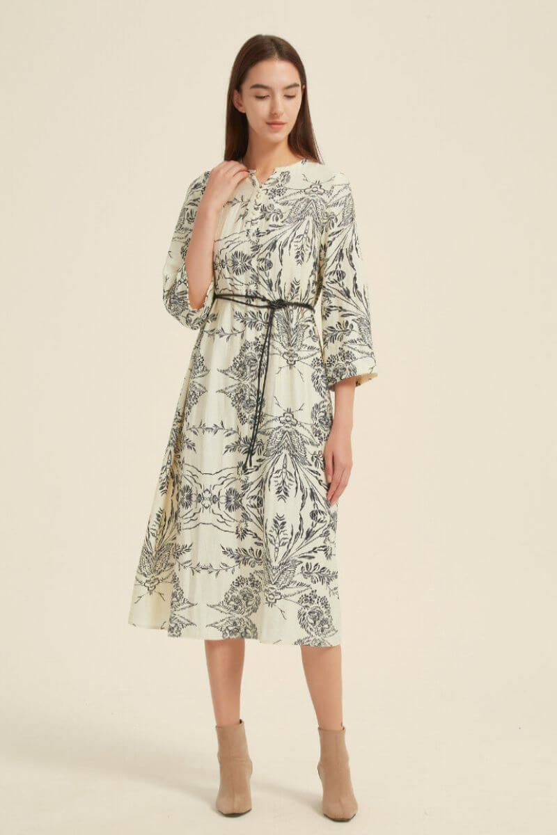 Full front studio view of a cream midi dress adorned with black floral patterns, finished with a skinny belt to define the waist.