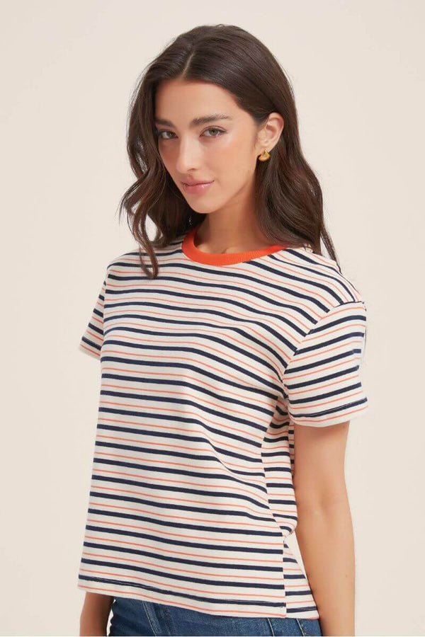 Side view of a woman wearing a cream short-sleeve T-shirt with navy and orange horizontal stripes and an orange ribbed neckline, styled with gold earrings and casual denim jeans.