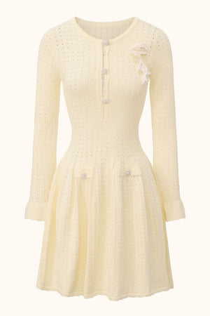 A cream pointelle knit long-sleeve mini dress with a fitted waist, flared skirt, decorative pearl buttons and a soft knitted flower brooch on the chest, photographed on a neutral background.