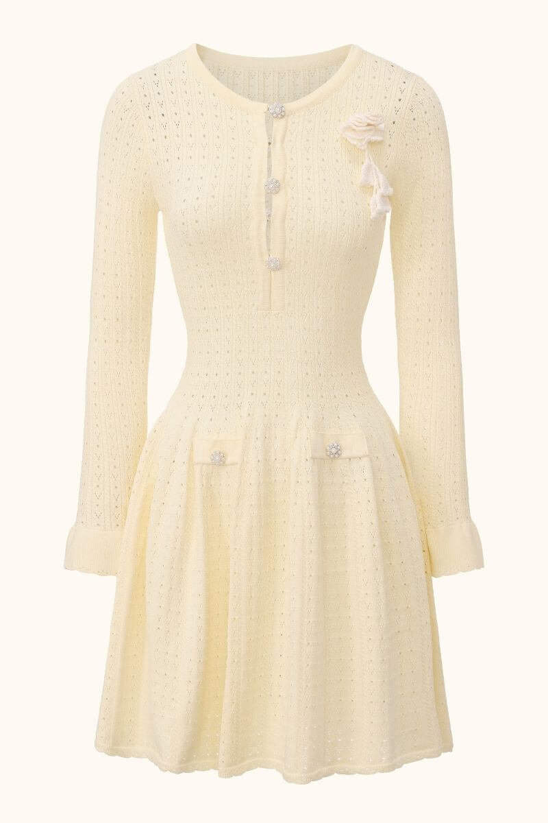 A cream pointelle knit long-sleeve mini dress with a fitted waist, flared skirt, decorative pearl buttons and a soft knitted flower brooch on the chest, photographed on a neutral background.