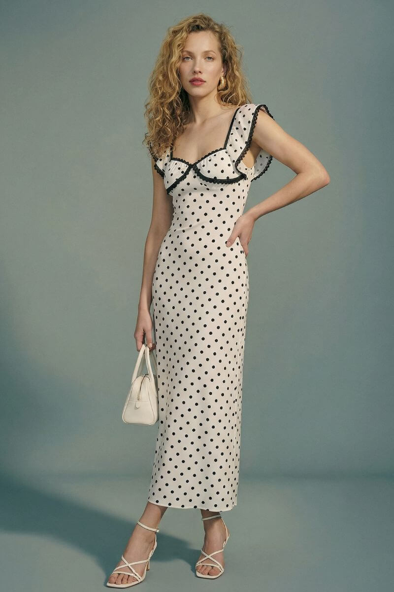 Model in a cream polka dot midi dress with black lace trim and flutter sleeves, captured at an angle to highlight the silhouette and delicate contrast details.