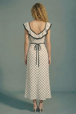 Model shown from the back wearing a cream polka dot midi dress with black lace trim, flutter sleeves, an open V back with adjustable tie detail, and white strappy heels.