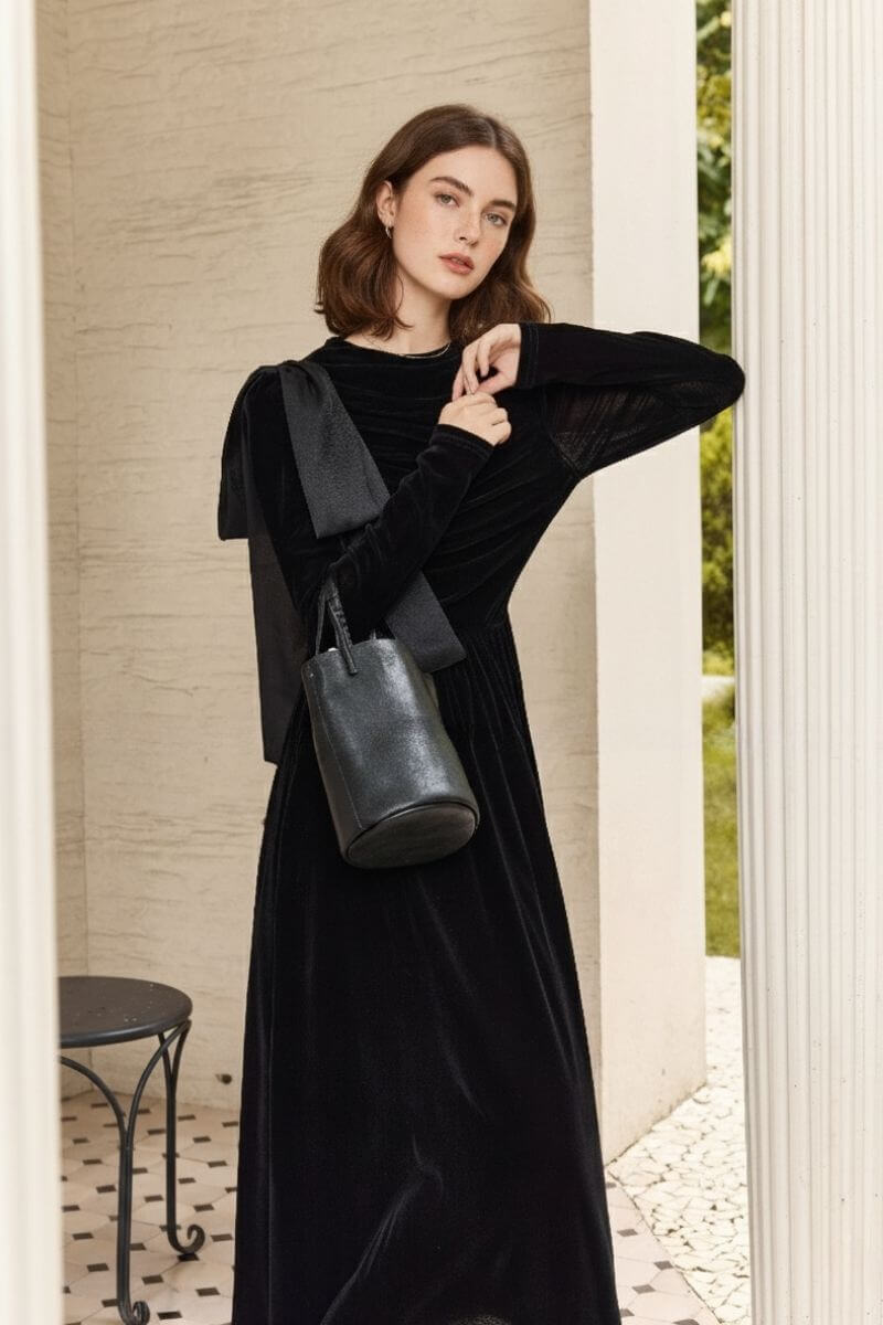 Model wearing a ruched black velvet long-sleeve dress with an oversized bow on the shoulder and carrying a black leather bag, standing beside a white column.