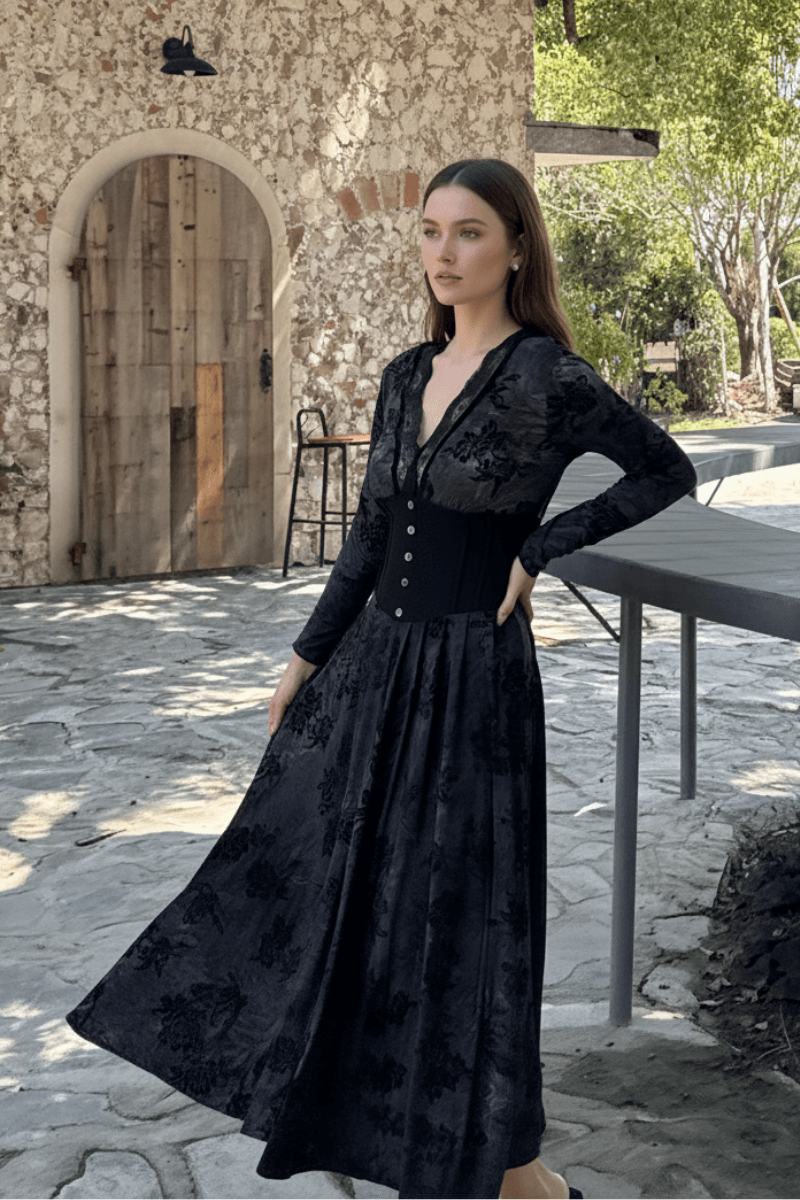 Model wearing a long black velvet maxi dress with floral patterns and corset waist detail, standing in a rustic stone courtyard with a wooden arched door in the background.