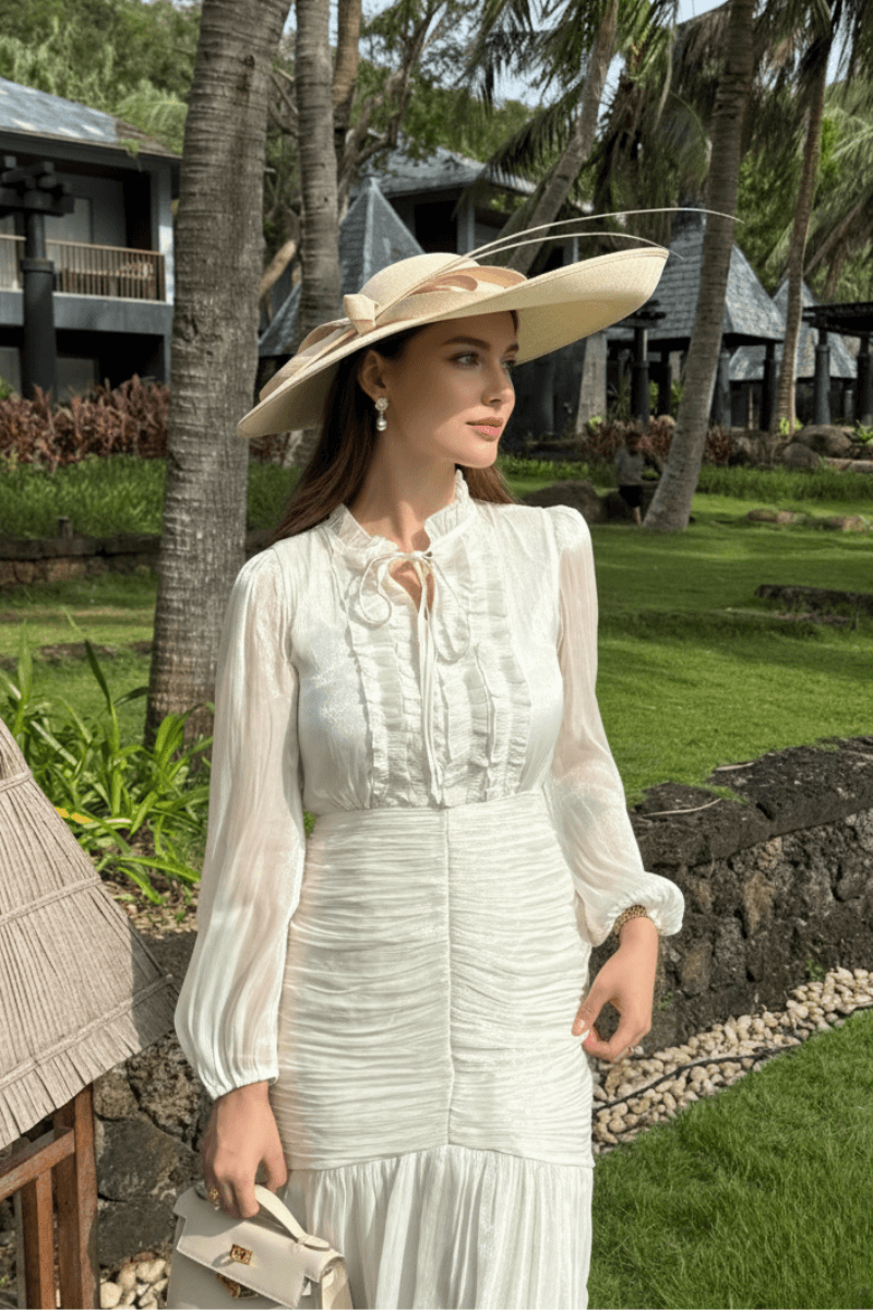 Close-up of a woman in a white ruffled maxi dress, accessorized with pearl earrings and a wide-brimmed hat, standing in a lush garden.