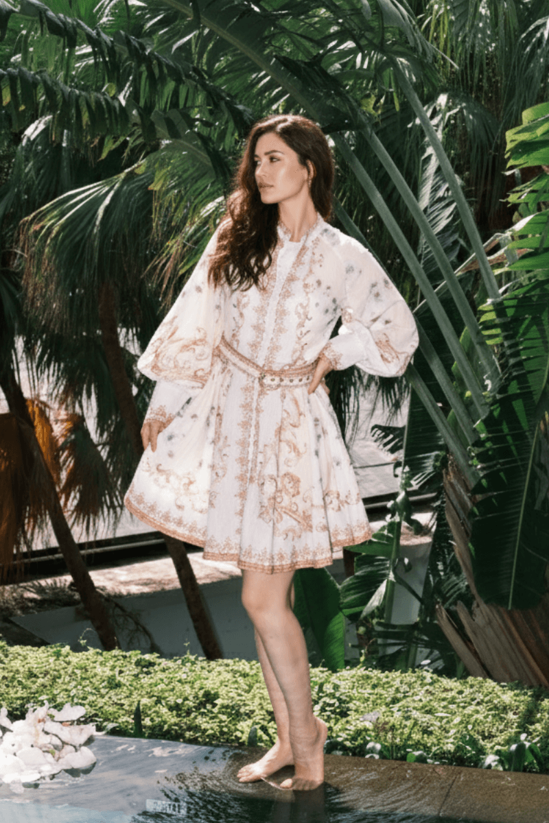 Woman wearing a white mini dress with intricate gold baroque print, belted waist, and flowy long sleeves, posing outdoors among tropical palm trees.