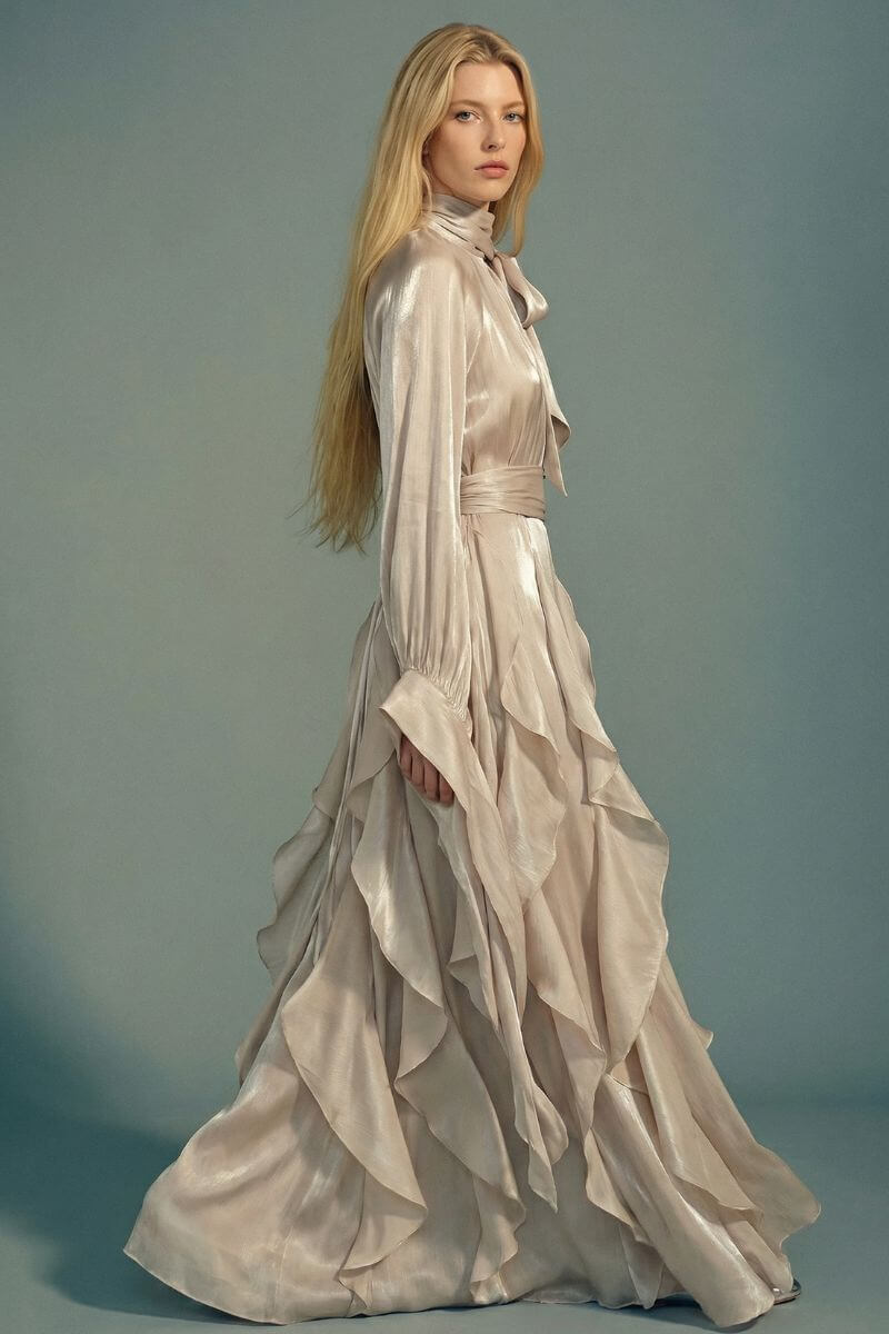 Side view of a silver satin maxi dress with long sleeves, bow neckline, and layered ruffles, captured mid-step to highlight the fabric’s soft sheen and fluid drape.