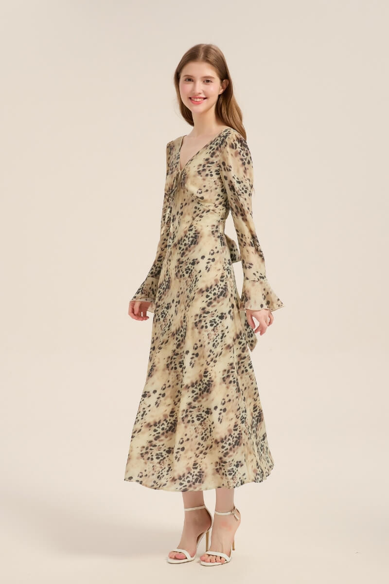Model smiling in a leopard print midi dress with soft drape, long sleeves, and tie-front neckline, paired with ankle-strap heels.