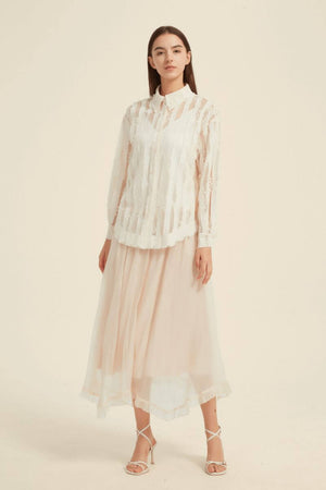 Front view of woman in ivory sheer lace stripe button-up blouse styled with blush pink midi skirt and strappy heels, minimal studio backdrop.