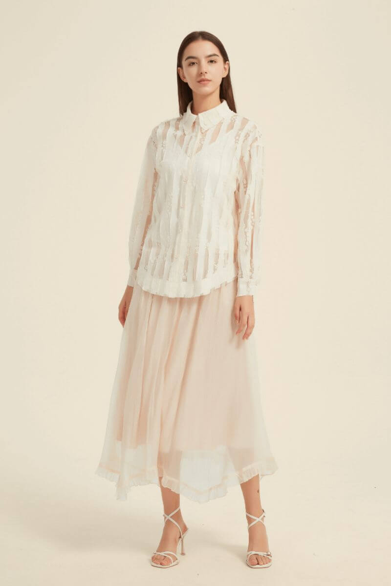 Front view of woman in ivory sheer lace stripe button-up blouse styled with blush pink midi skirt and strappy heels, minimal studio backdrop.
