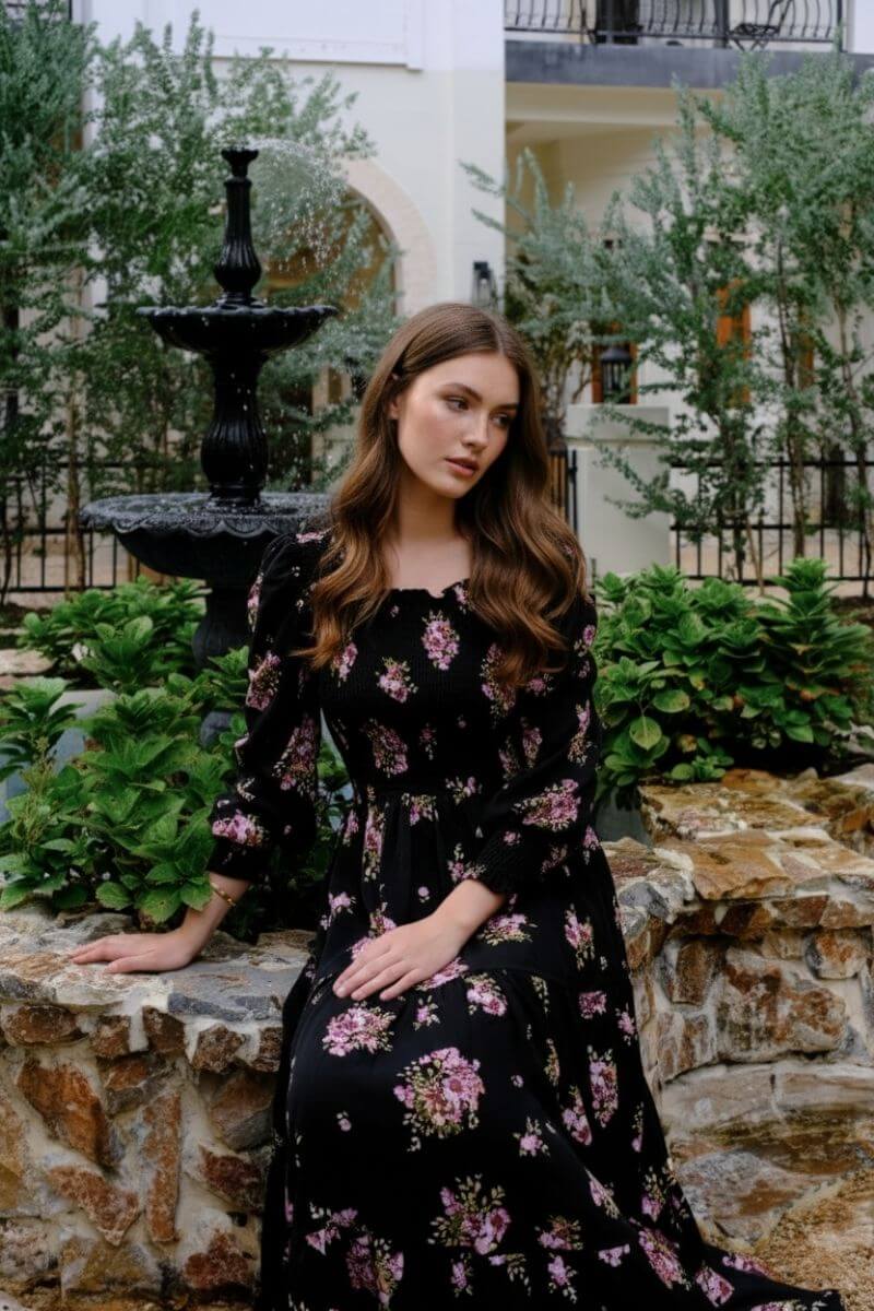 Model seated beside a black fountain wearing a black smocked maxi dress with pink floral motifs and tiered skirt, surrounded by green foliage and stone garden walls for a romantic countryside feel.