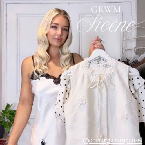 Woman preparing outfits in a soft white bedroom, holding a lace and polka-dot dress from Sioéne — GRWM fashion moment with feminine elegance.