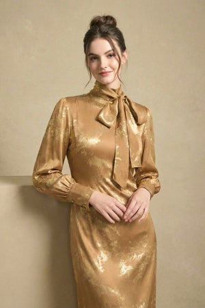 Woman smiling in a studio wearing a gold floral satin bow-tie dress with long sleeves.
