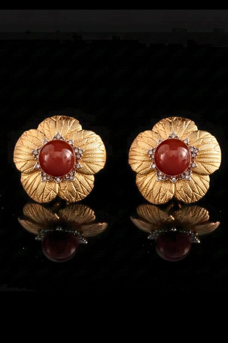 Two gold flower-shaped stud earrings featuring a domed red carnelian center and fine petal textures, arranged side by side on a black reflective background.