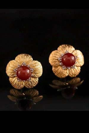 A pair of gold floral stud earrings with textured petal detailing and a polished red carnelian center, photographed on a glossy black surface.