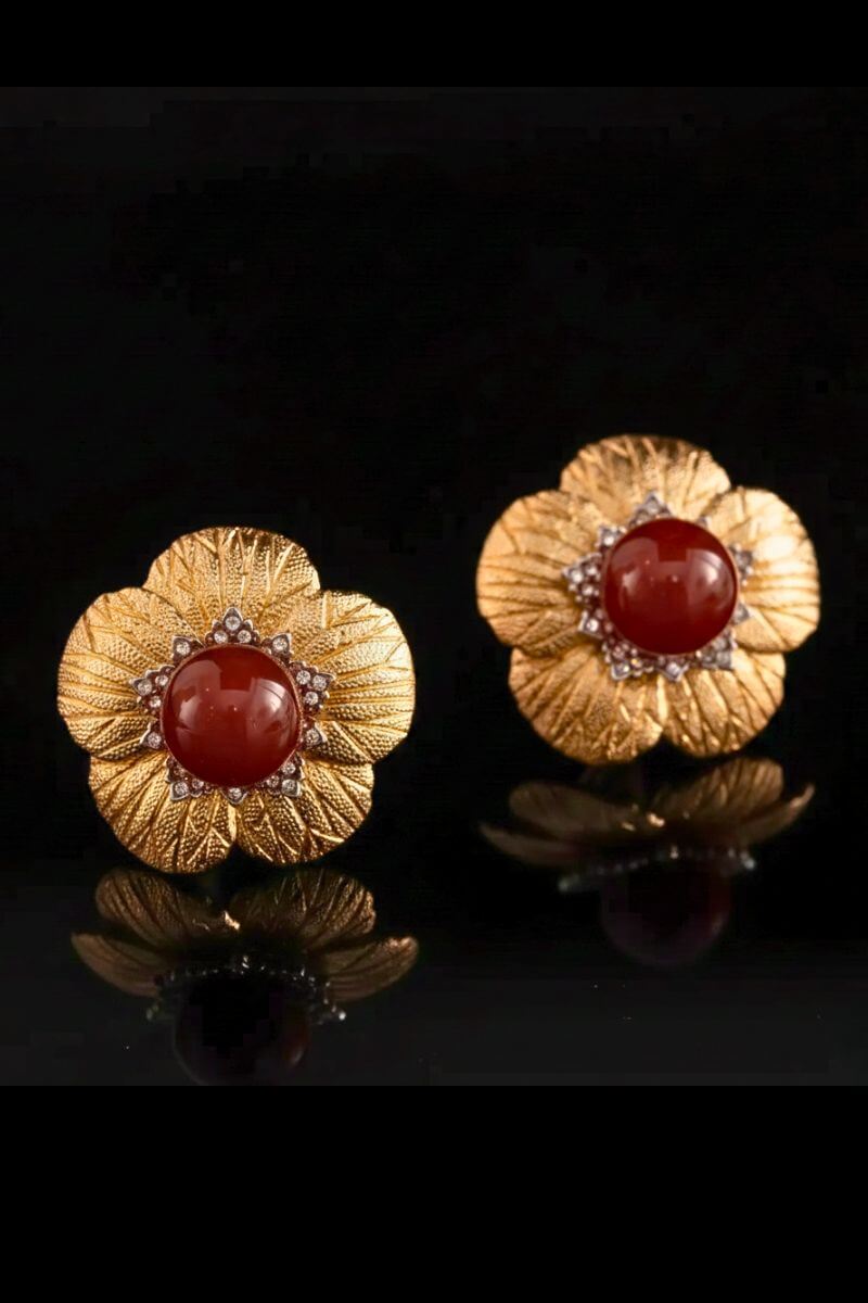 A pair of gold floral stud earrings with textured petal detailing and a polished red carnelian center, photographed on a glossy black surface.